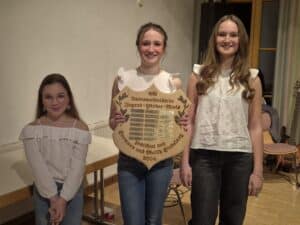 Drei Mädchen stehen nebeneinander und lächeln in die Kamera. Das mittlere hält eine Holztafel mit der Aufschrift "SV Untermaiselstein Jugend-Förder-Preis" und Jahresplaketten in der Hand.