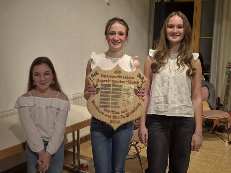 Drei Mädchen stehen nebeneinander und lächeln in die Kamera. Das mittlere hält eine Holztafel mit der Aufschrift "SV Untermaiselstein Jugend-Förder-Preis" und Jahresplaketten in der Hand.